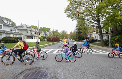 Children bike through intersection following adult as another adult pauses car traffic from other direction