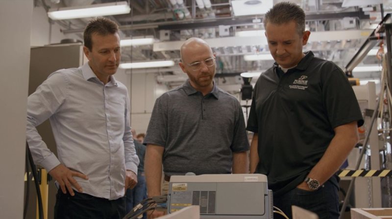 Three men examine machine measuring electromagnetic performance of coils.